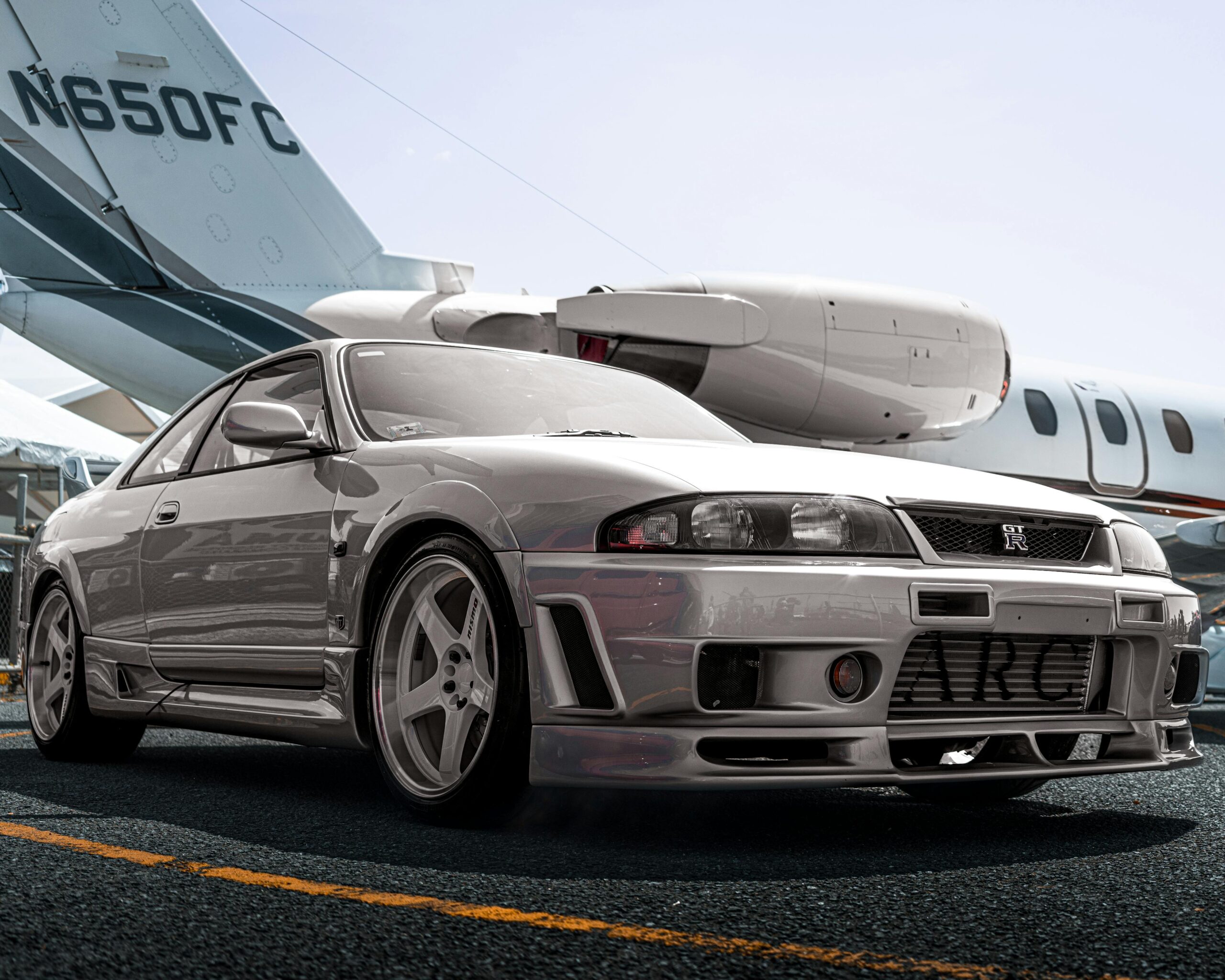 A silver sports car parked on tarmac with a private jet in the background in an urban setting.